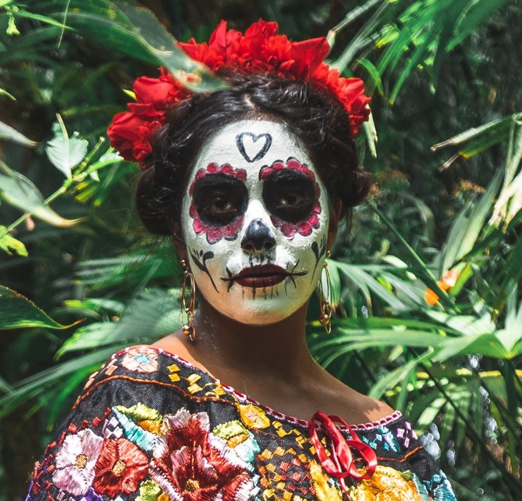 standing woman with painted face surrounded by green plants